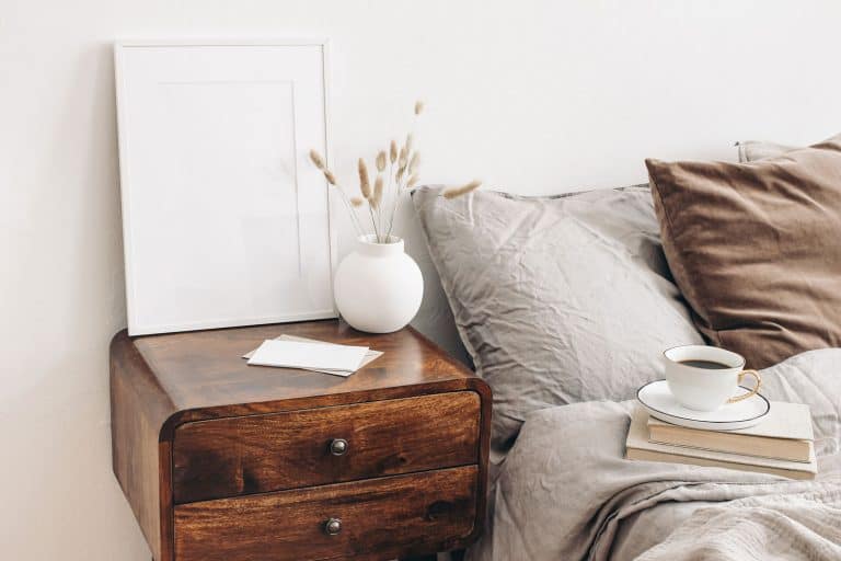 A small wooden nightstand placed next to a gray bed with gray and brown pillows, How Far Should Bedside Table Be From Bed?