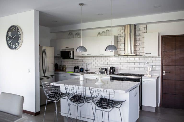 Open space apartment room with white painted walls, white kitchen island with stainless steel chairs and dangling lamps, How Wide Should A Kitchen Island Be?