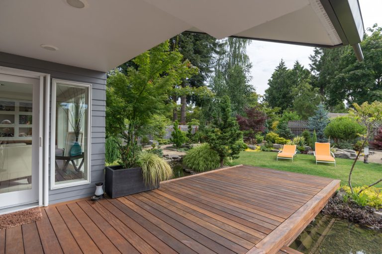 A perspective view of a contemporary Pacific Northwest home with a deck bridging a pond that leads to a pair of modern yellow loungers in a landscaped yard, Can You Sand A Deck After It Rains?